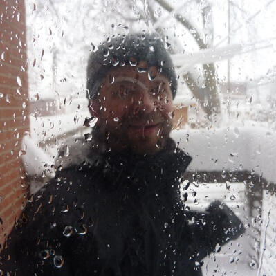 A man in a knit cap standing on a snowy porch through glass that is obscured by drops of water.